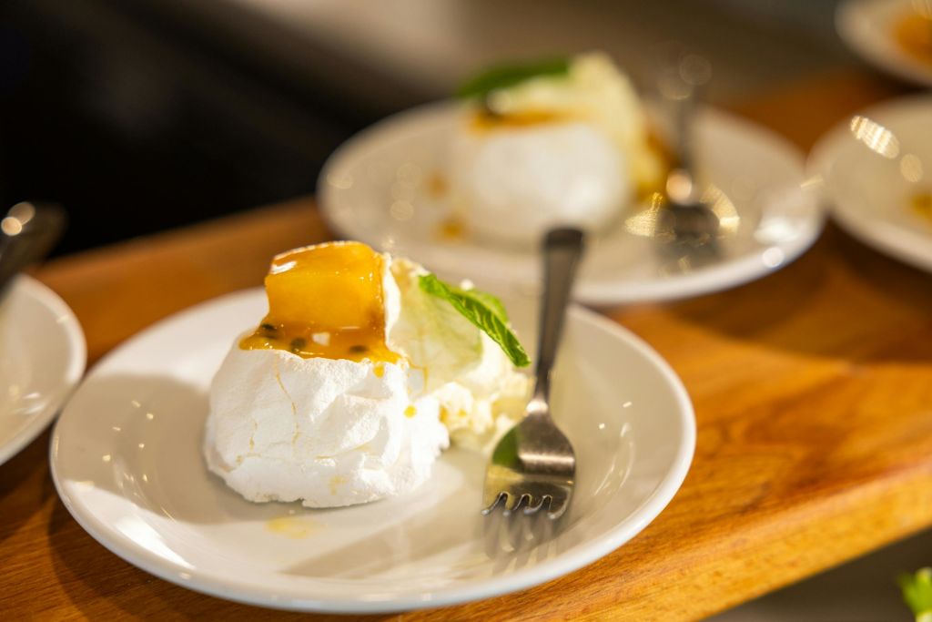 휘핑 크림, 열정 과일 소스, 민트 잎을 얹은 파블로바 두 접시. 각 접시에는 포크가 놓여 있습니다.