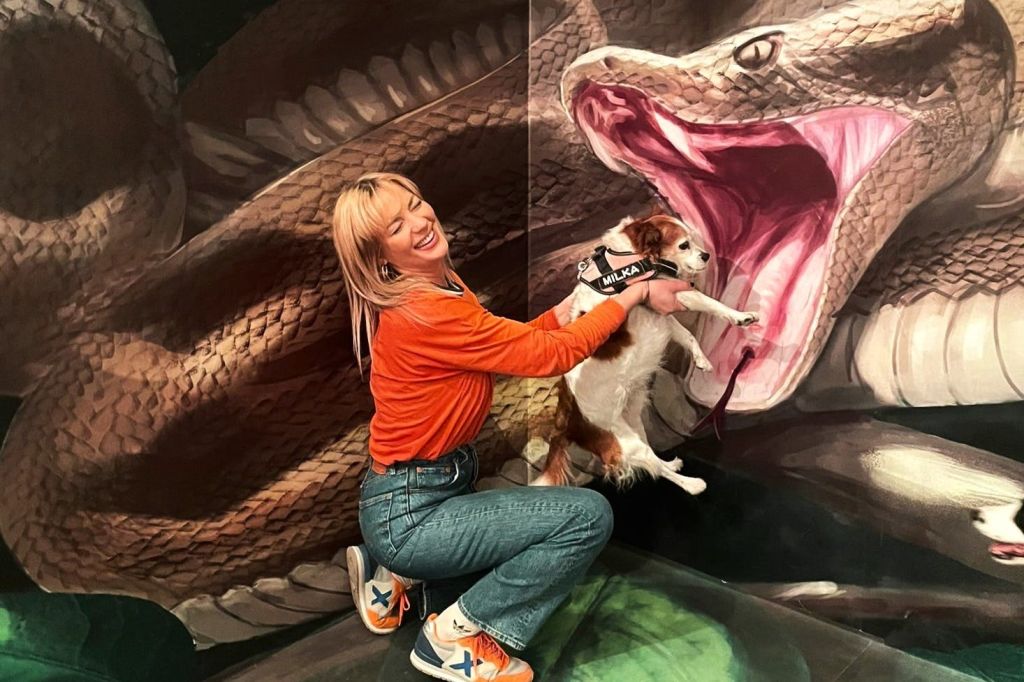 주황색 셔츠를 입은 한 여성이 입을 벌린 채 커다란 뱀 그림 앞에서 작은 개와 함께 포즈를 취하고 있습니다.