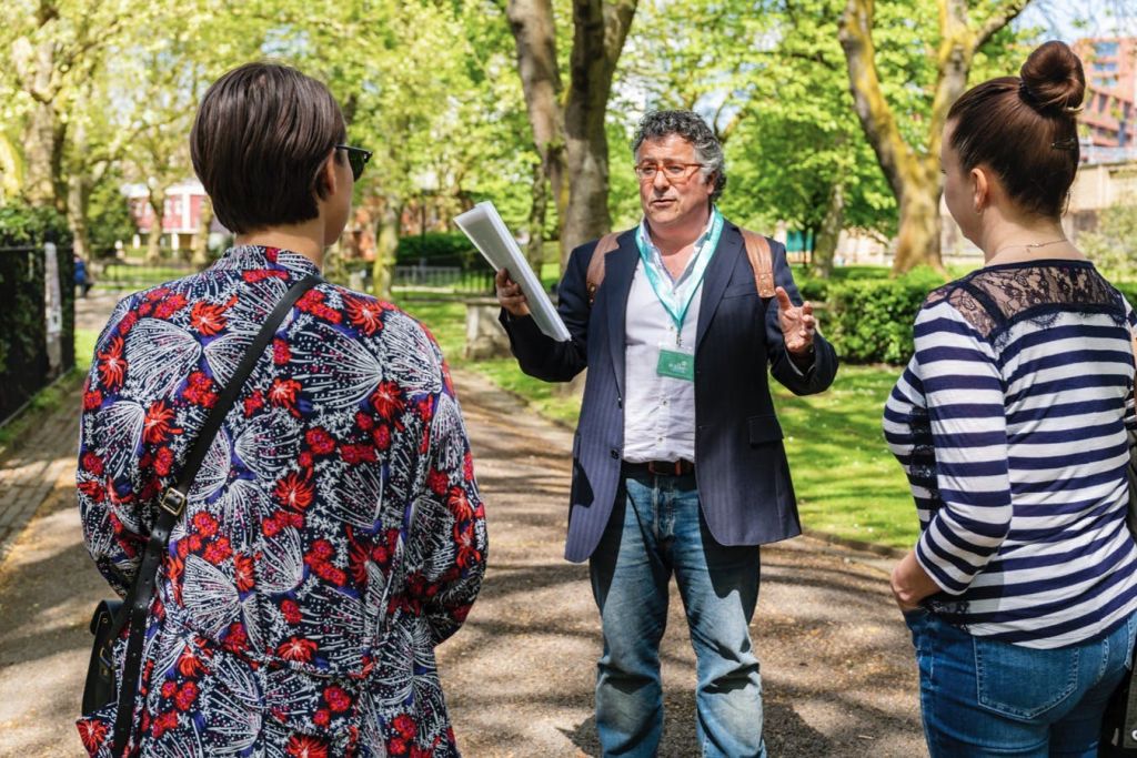 공원에서 나무가 있고 햇살이 내리쬐는 초록색 배경의 두 여성과 대화를 나누는 동안 폴더가 있는 남성이 제스처를 취하고 있습니다.