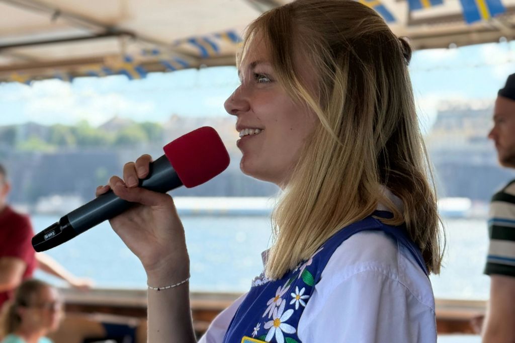 빨간색 폼 커버가 달린 마이크를 들고 미소를 짓고 물과 흐릿한 배경이 있는 보트에서 말하는 여성.