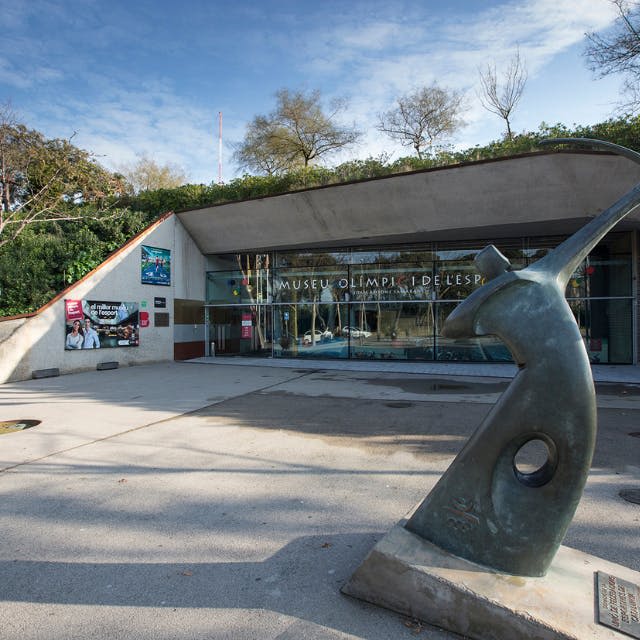 커다란 창문, 곡선형 청동 조형물, 나무를 배경으로 한 현대적인 건물 입구. "Museu Olímpic i de l 'Esport" 라는 표지판이 있습니다.