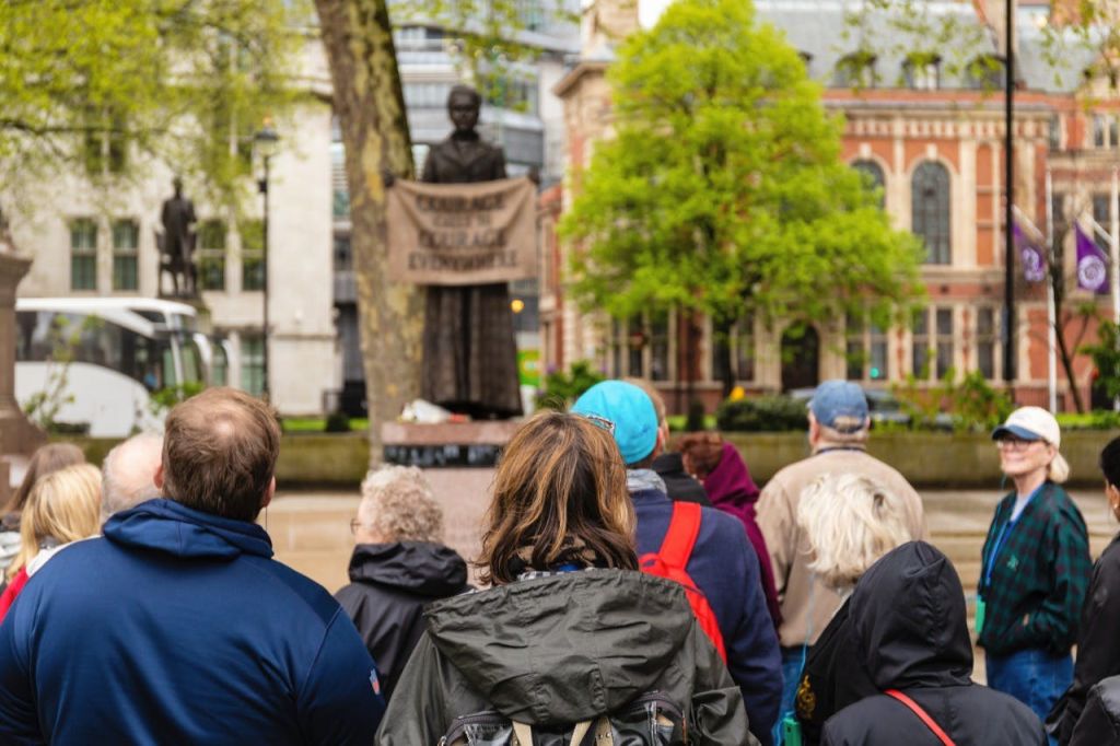 나무가 우거진 야외 광장과 역사적인 건물을 배경으로 한 무리의 사람들이 배너가 달린 동상을 마주하고 서 있습니다.