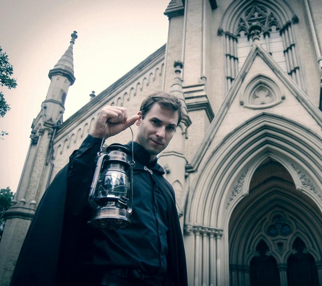 아치형 창문과 화려한 디테일이 돋보이는 고딕 양식의 건물 앞에 랜턴을 들고 있는 남성.