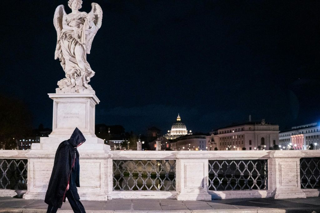 어두운 망토를 입은 사람이 밤에 다리를 걷고 있는데, 조각상과 조명 돔을 배경으로 하고 있습니다.