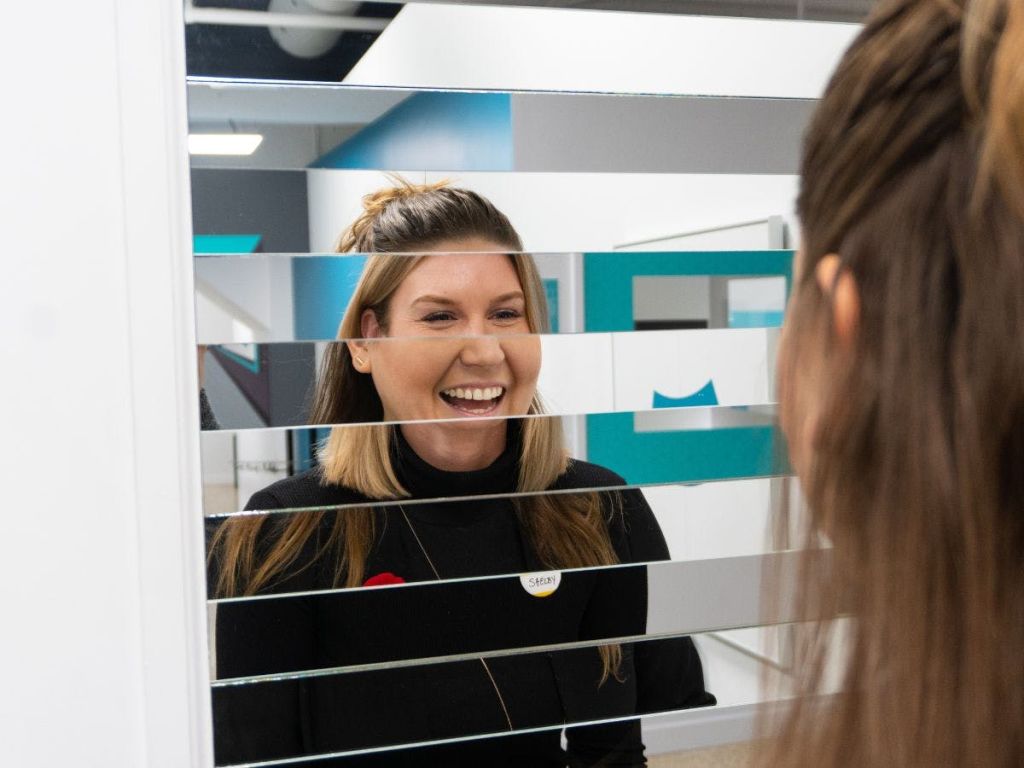 부분적으로 덮인 유리 칸막이 뒤에 "Sarah" 이름 배지를 달고 다른 사람과 상호 작용하는 미소 짓는 여성.