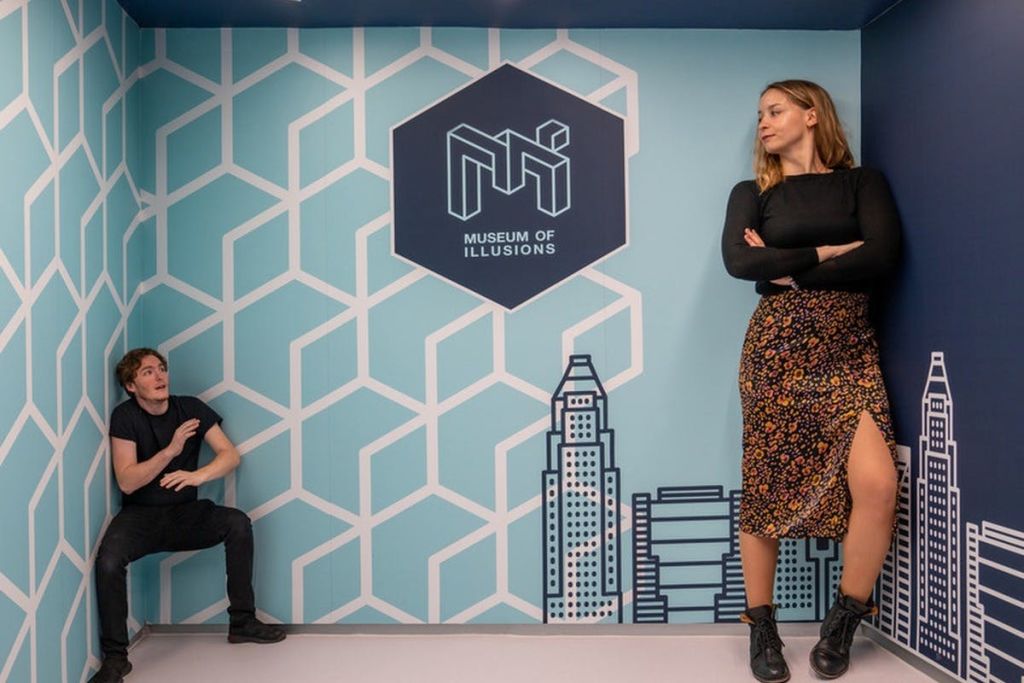기하학적 벽 디자인과 "환상 박물관" 로고가 있는 방에 두 사람이 있습니다. 하나는 원근감으로 인해 더 크게 보입니다.