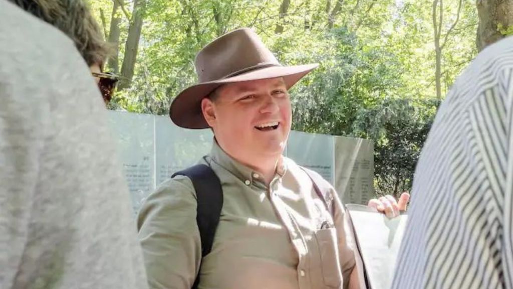 활짝 핀 모자를 쓰고 나무와 유리 패널을 배경으로 배낭을 메고 야외에서 웃고 있는 남성.