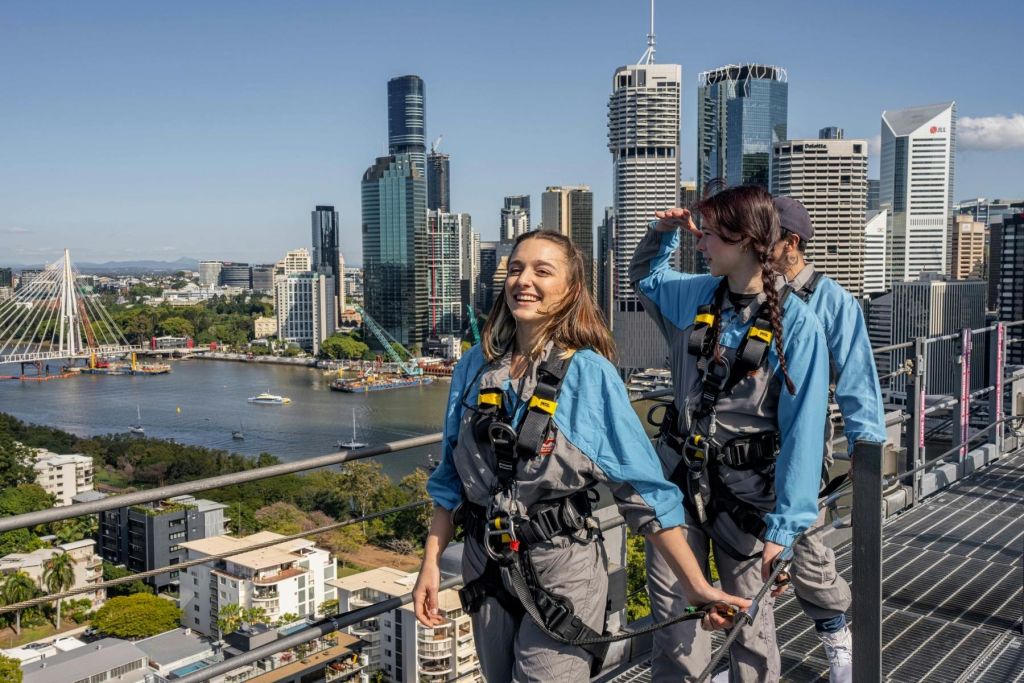 도심 옥상에는 고층빌딩과 강을 배경으로 세 사람이 안전 장비를 착용하고 있습니다. 한 사람은 웃고 있고, 다른 한 사람은 눈을 감고 있습니다.