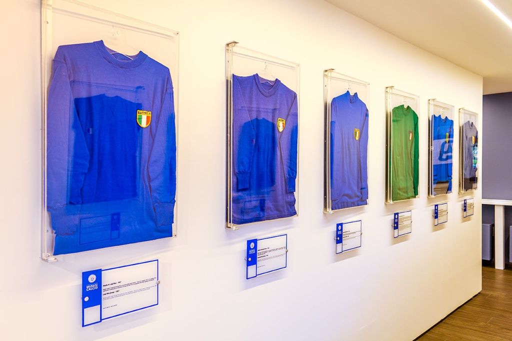 파란색 3개와 녹색 1개를 포함한 네 개의 프레임 빈티지 축구 유니폼이 벽에 표시되고 그 아래에 설명 플래카드가 있습니다.