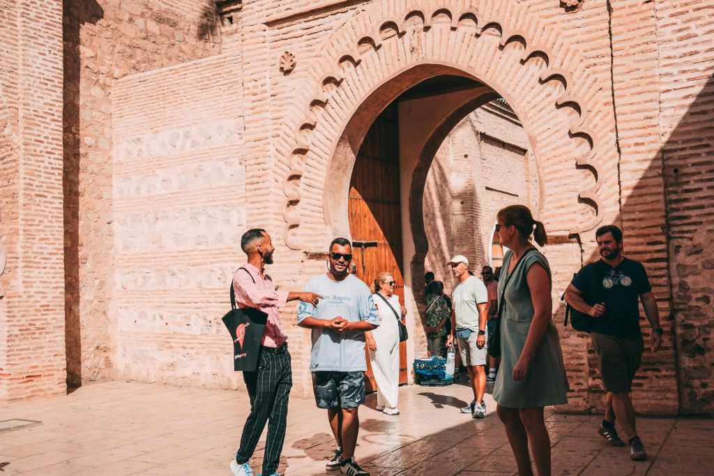 화창한 날, 커다랗고 화려한 석조 아치길 근처에서 서로 교류하며 걷는 사람들. 일부는 가방을 들고 캐주얼한 옷을 입고 있습니다.