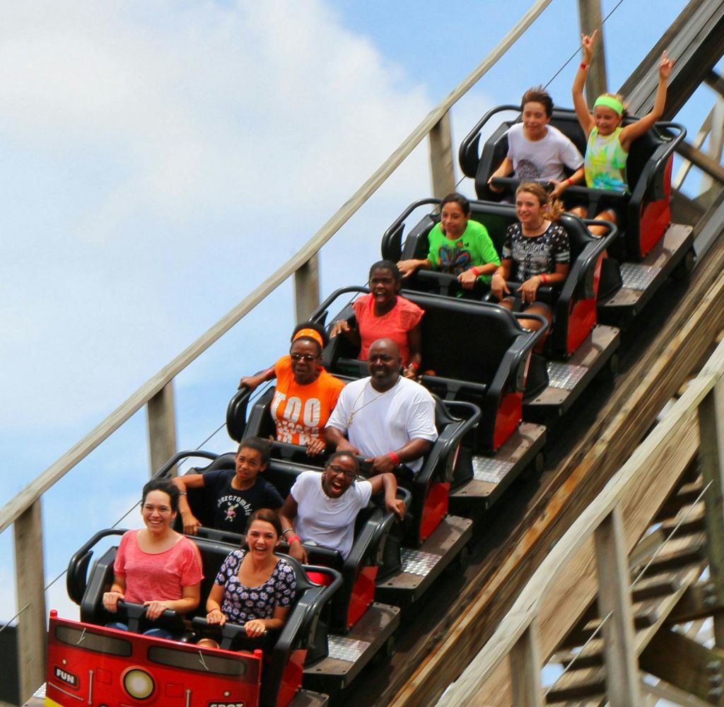 빨간색 롤러코스터를 탄 9명이 나무 트랙을 내려가며 미소를 짓고 푸른 하늘을 향해 팔을 들어 올립니다.