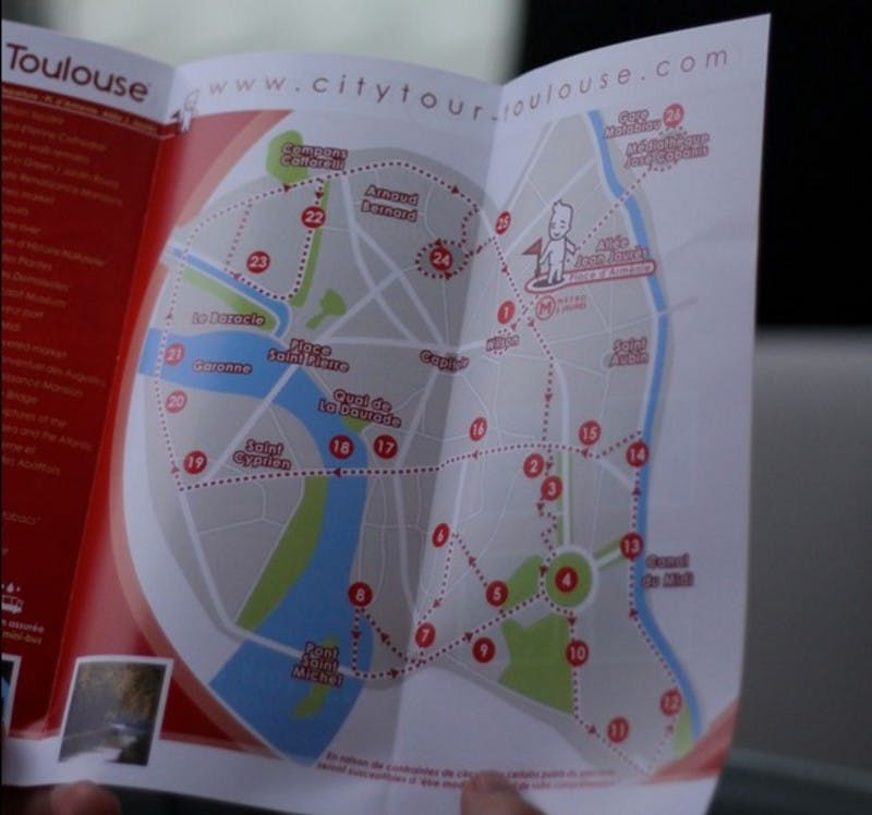 파란색과 녹색 영역을 배경으로 빨간색으로 번호가 매겨진 명소와 경로를 보여주는 툴루즈 시티 투어 지도를 들고 있는 사람.