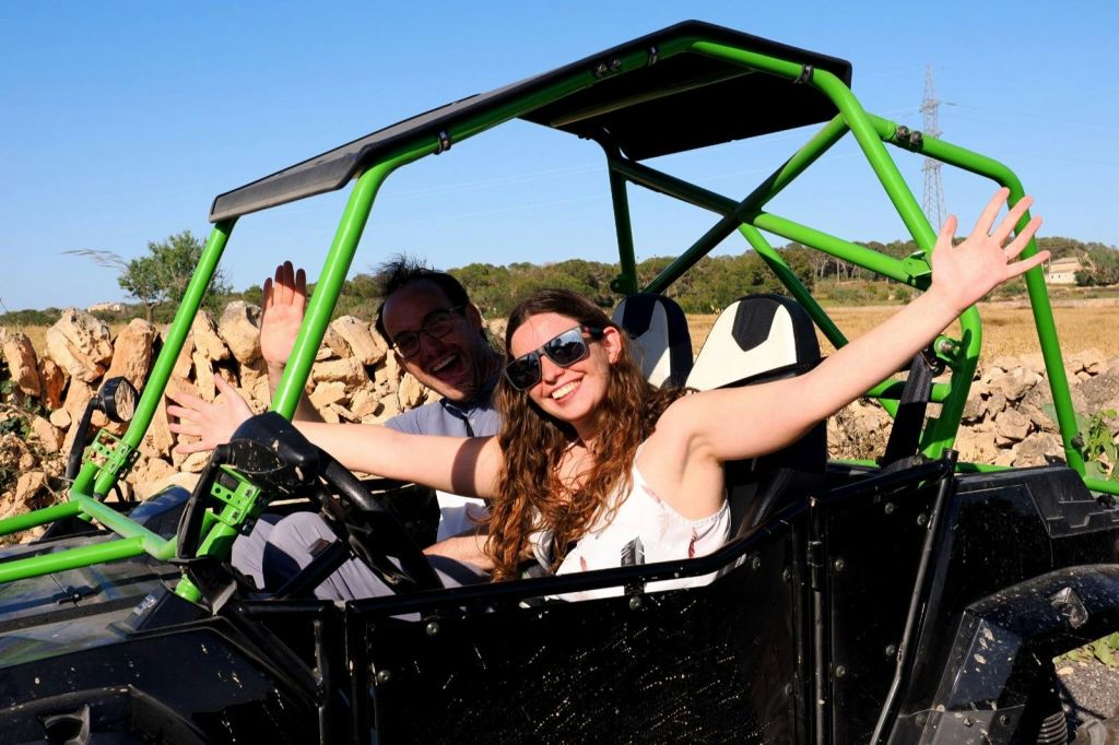선글라스를 쓴 두 사람이 맑고 푸른 하늘 아래 초록색과 검은색 오프로드 차량 안에서 두 팔을 들고 포즈를 취하고 있습니다.