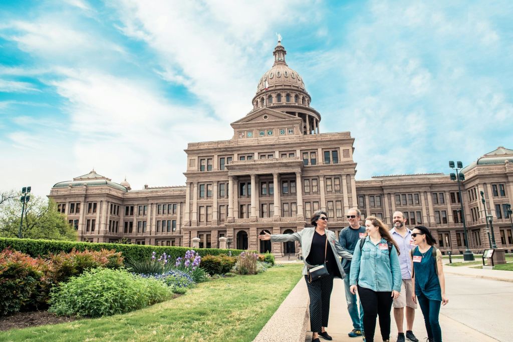 밝고 푸른 하늘 아래 돔이 있는 유서 깊은 대형 건물 앞에서 한 무리의 사람들이 걷고 이야기하고 있습니다.