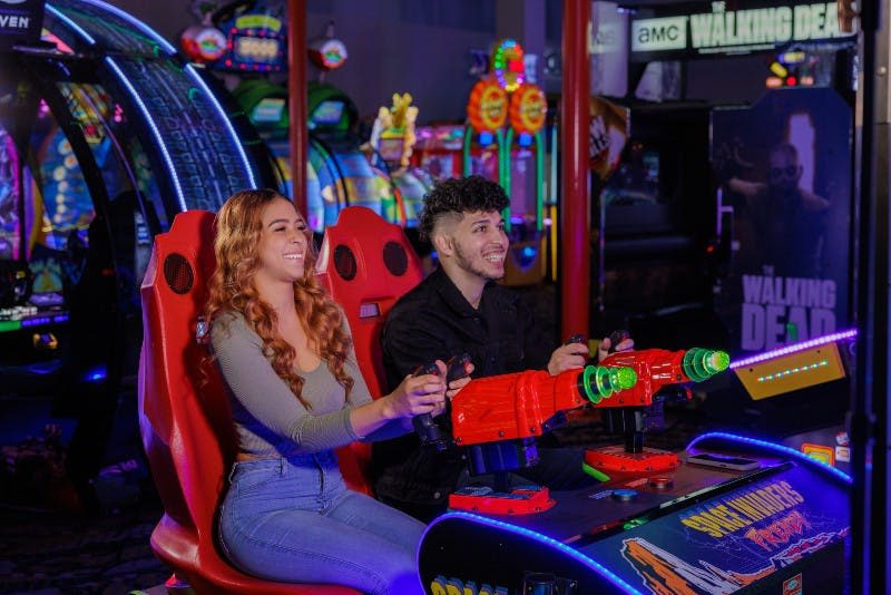 화려한 아케이드 기계로 둘러싸인 빨간색과 녹색 총으로 아케이드 게임을 하면서 웃고 있는 두 사람.