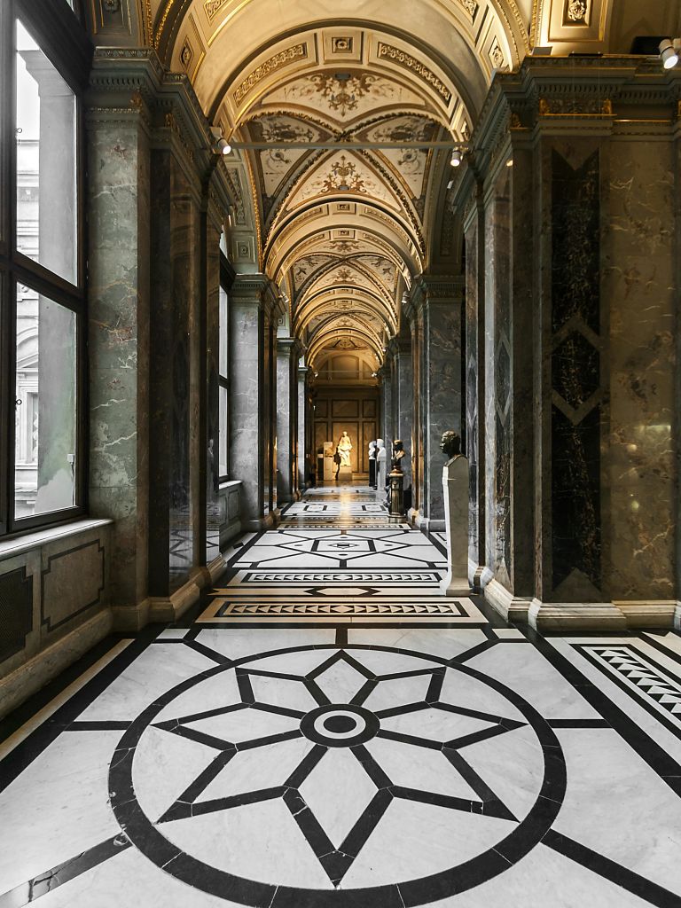 화려한 천장 디자인과 동상, 정교한 바닥 패턴이 있는 웅장한 대리석 복도로, 커다란 창문에서 자연 채광이 들어옵니다.
