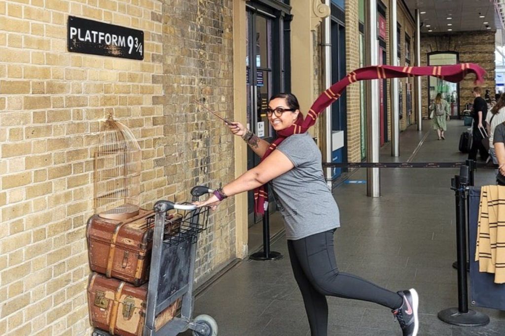 한 사람이 수하물 트롤리 옆에 있는 플랫폼 9 3/4 어트랙션에서 봉과 스카프를 들고 열정적으로 포즈를 취하고 있습니다.