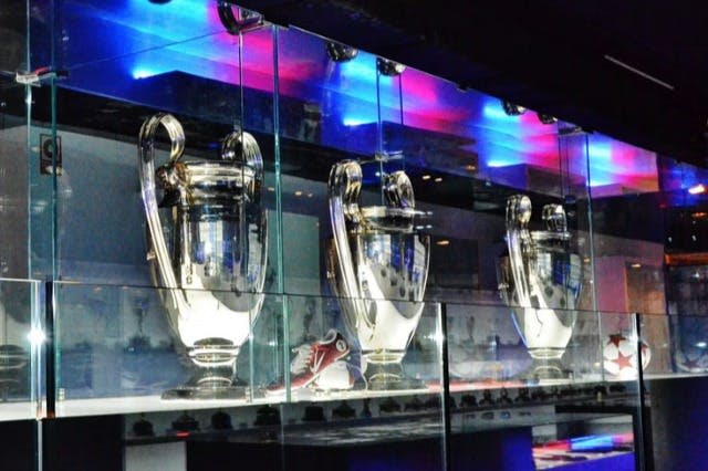 유리 선반에 세 개의 커다란 은색 트로피가 진열되어 있으며, 화려한 조명이 유리에 반사됩니다. 개체는 배경에 있습니다.