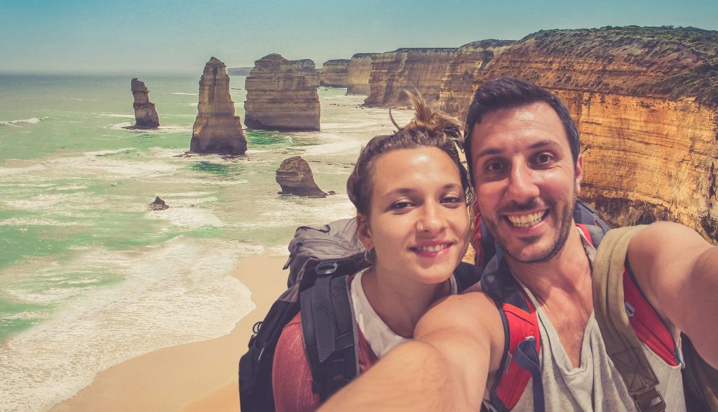 높고 겹겹이 쌓인 절벽이 있는 험준한 해안선 앞에 서서 웃고 있는 커플이 배낭을 메고 셀카를 찍고 있습니다.