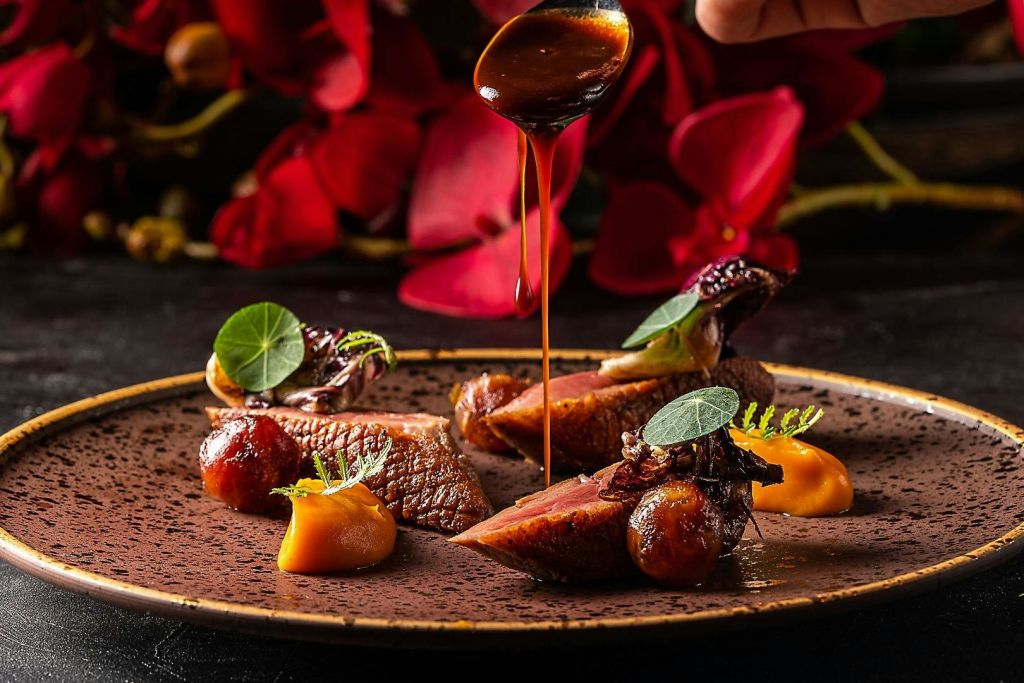 구운 고기, 구운 야채, 소스, 장식이 들어간 고급 요리로, 숟가락으로 튀기는 소스와 화사한 붉은 꽃을 배경으로 합니다.