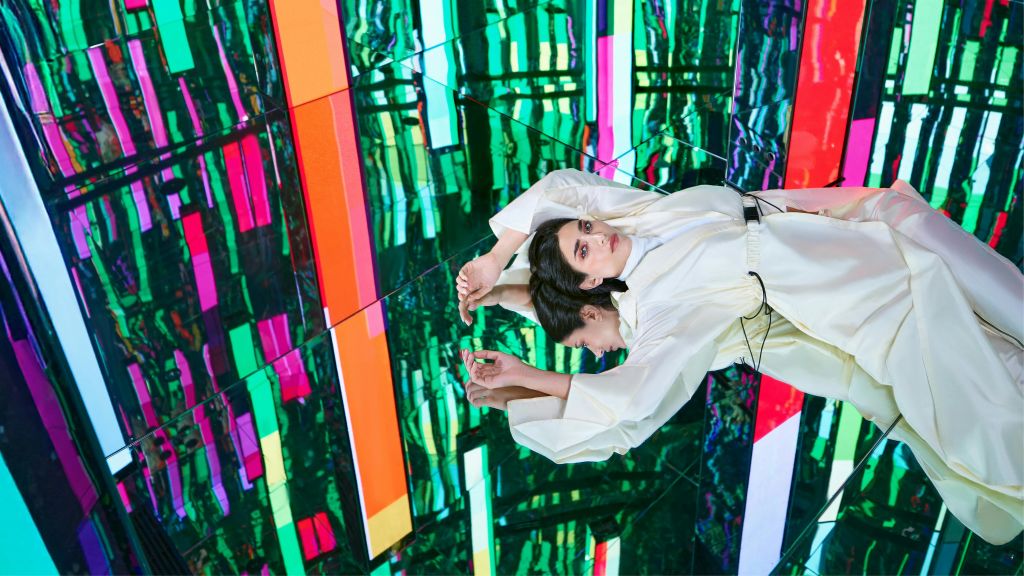 흰색 의상을 입은 여성이 배경에 화려하고 추상적인 조명 패턴이 있는 반사 표면에 누워 있습니다.
