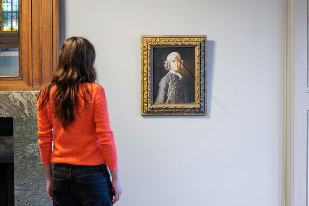 주황색 스웨터를 입은 여성이 얼굴에 붉은 페인트가 흩뿌려진 역사적 인물의 액자 초상화를 바라보고 있습니다.