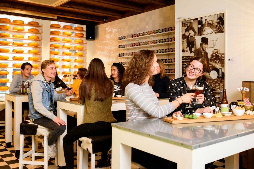 치즈 바퀴 선반과 통조림을 배경으로 아늑한 치즈 가게 내부의 흰색 테이블에 앉아 있는 사람들.