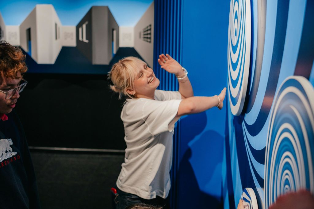 흰색 셔츠를 입은 사람이 동심원 무늬가 있는 파란색 벽에 기대어 미소를 짓고 있습니다. 기하학적 도형이 배경에 표시됩니다.