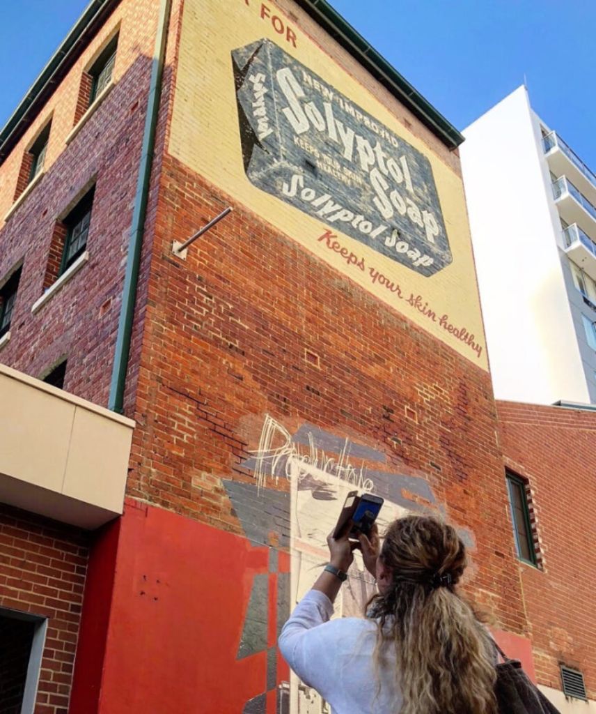 한 사람이 Solvol 비누를 광고하는 벽돌 건물에서 벽화 사진을 찍고 있습니다. 현대식 건물이 배경으로 보입니다.