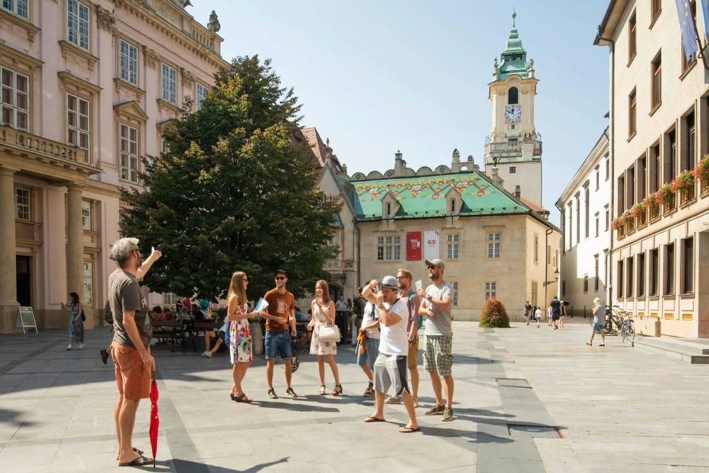 유서 깊은 건물과 시계탑을 배경으로 햇살이 내리쬐는 광장에서 한 무리의 사람들이 서서 이야기를 나누고 있습니다.