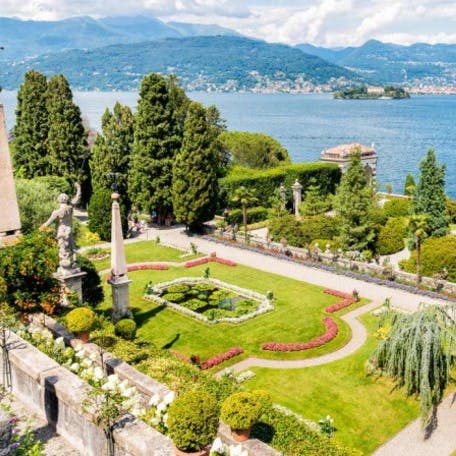 산을 배경으로 조각상, 잘 관리된 울타리, 길, 활기찬 화단이 있는 아름답게 조경된 호숫가 정원입니다.