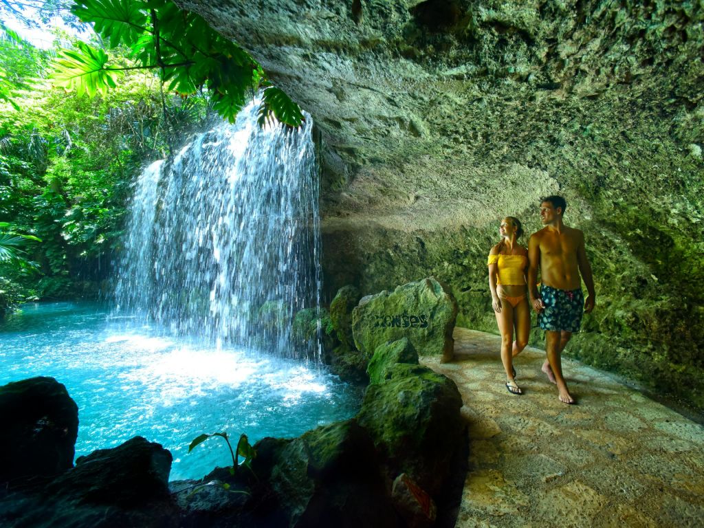 한 커플이 동굴 안쪽 폭포 근처의 돌길을 걷고 있는데, 머리 위로 녹음이 우거진 풍경이 보입니다.