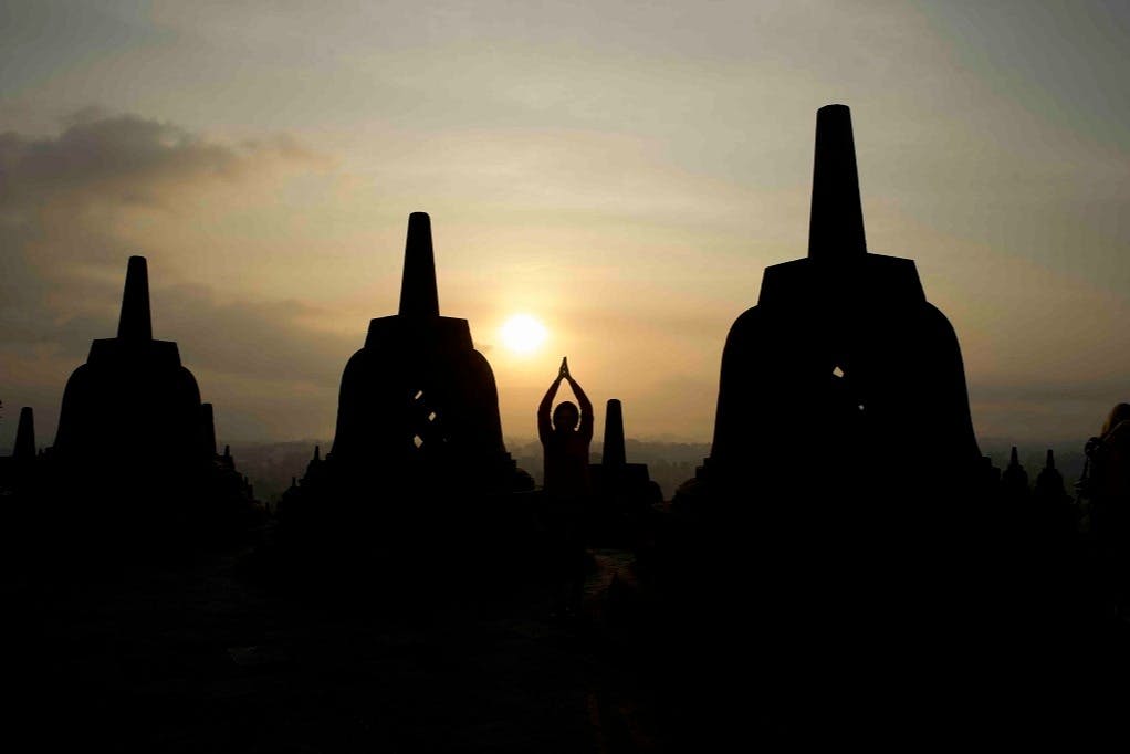 해가 뜰 무렵, 커다란 종 모양의 스투파에 둘러싸인 채 손을 들고 기도하는 사람의 실루엣.