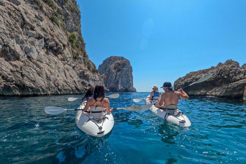 화창한 하늘 아래 바위 절벽 사이에 있는 맑고 푸른 물에서 카약을 타는 네 명의 사람들.