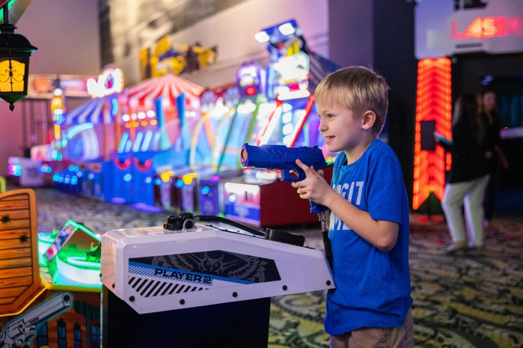 다채로운 기계로 둘러싸인 밝게 빛나는 게임 센터에서 레이저 슈팅 아케이드 게임을 하는 소년.