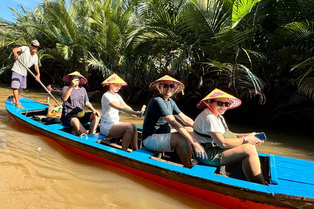 원뿔 모양의 모자와 선글라스를 쓴 세 사람이 파란색과 빨간색 보트에 앉아 열대 수로를 지나고 있습니다.