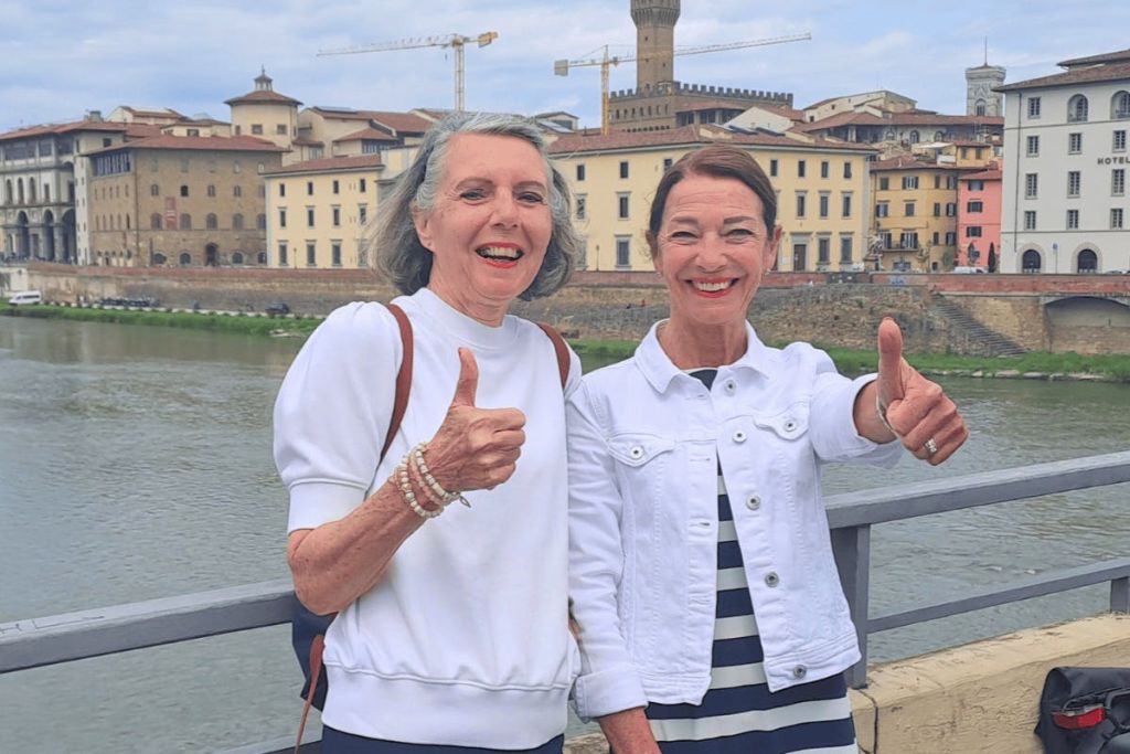 강변 마을과 건축 크레인을 배경으로 두 여성이 다리 위에 서서 미소를 짓고 엄지손가락을 치켜세우고 있습니다.