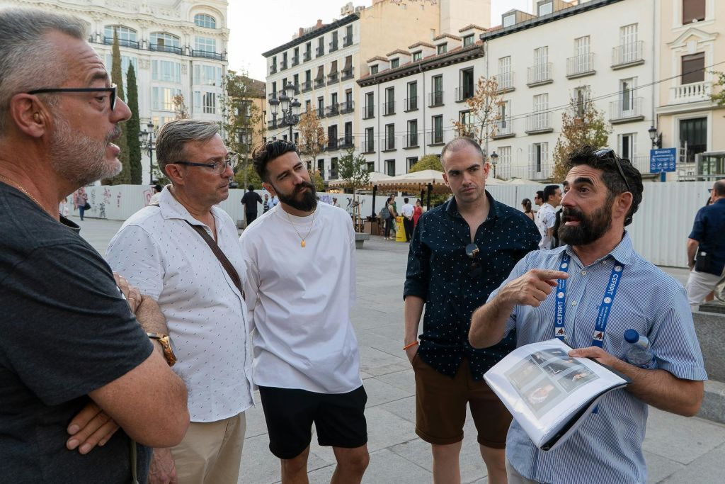 역사적인 건물로 둘러싸인 도시 광장에서 대화하는 네 명의 남성과 그 배경에 있는 몇 명의 사람들.