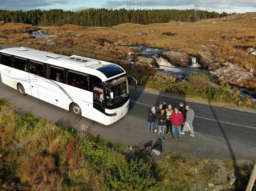 흰색 투어 버스 근처에 서 있는 한 무리의 사람들이 폭포와 잔디가 우거진 언덕이 있는 아름다운 강 옆의 좁은 도로에 주차되어 있습니다.