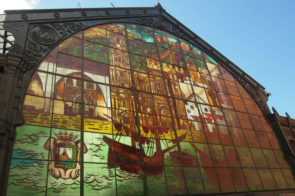 푸른 하늘을 배경으로 선명한 색상으로 건축 구조물, 선박 및 다양한 장면을 묘사한 대형 스테인드글라스 창문.