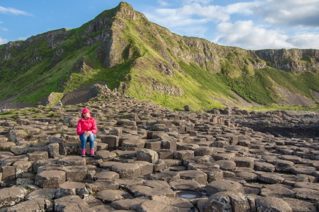 분홍색 재킷을 입은 사람이 부분적으로 흐린 하늘 아래 녹색 언덕을 배경으로 자이언트 코즈웨이에 앉아 있습니다.