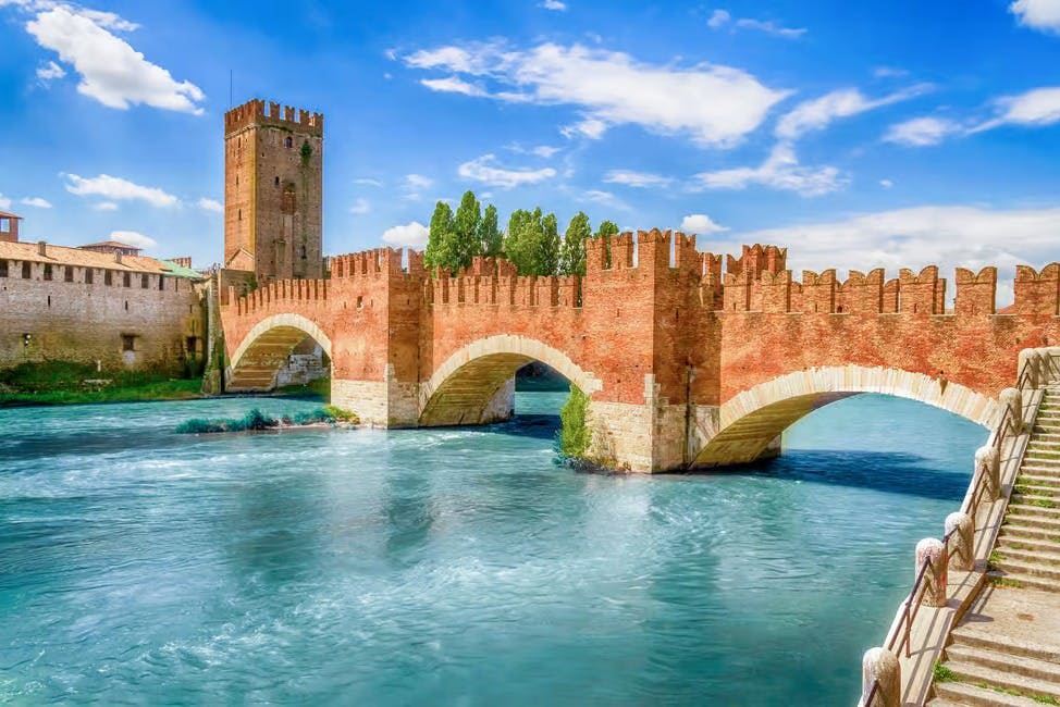 부분적으로 흐린 하늘을 배경으로 푸른 녹색 강 위에 아치가 있는 역사적인 붉은 벽돌 다리. 탑과 나무를 배경으로.