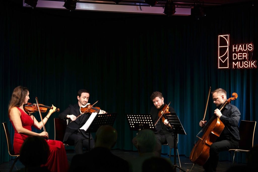 세 명의 뮤지션이 현악기로 무대에서 연주하며, 음악 스탠드가 있습니다. 청중은 세심하게 관찰합니다.