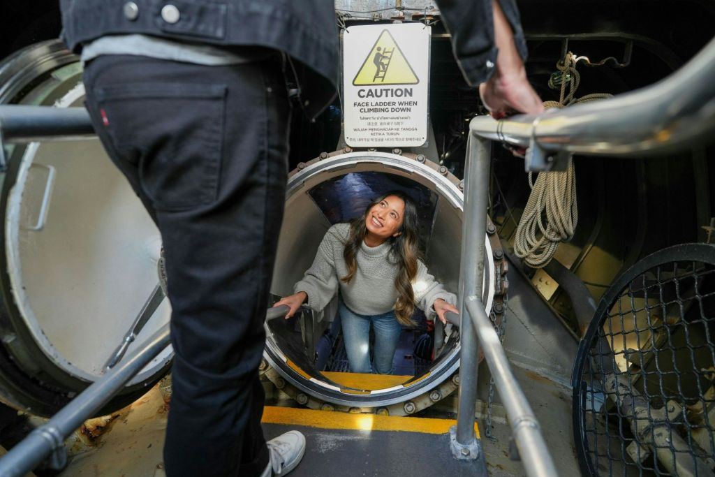 기계식 환경에서 원형 해치를 통해 올라가는 동안 웃고 있는 여성. 위에 주의 표지판이 보입니다.