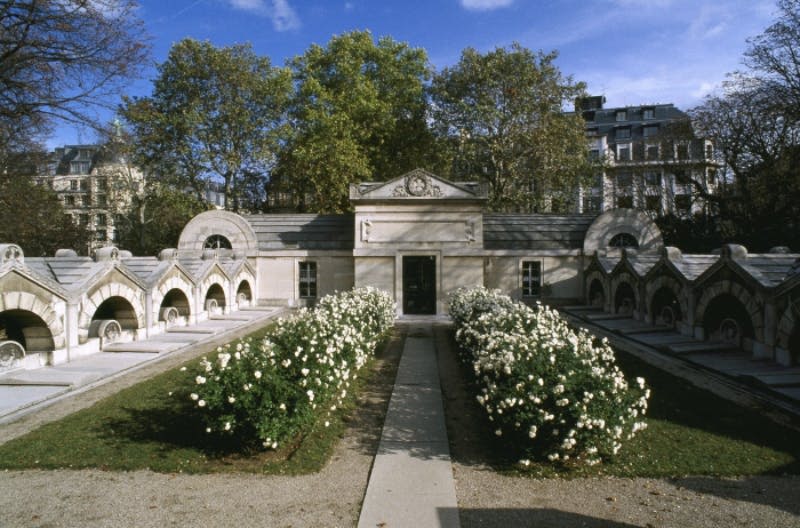 맑고 푸른 하늘 아래 하얀 꽃과 통로, 나무로 둘러싸인 아치형 무덤이 있는 석조 묘.