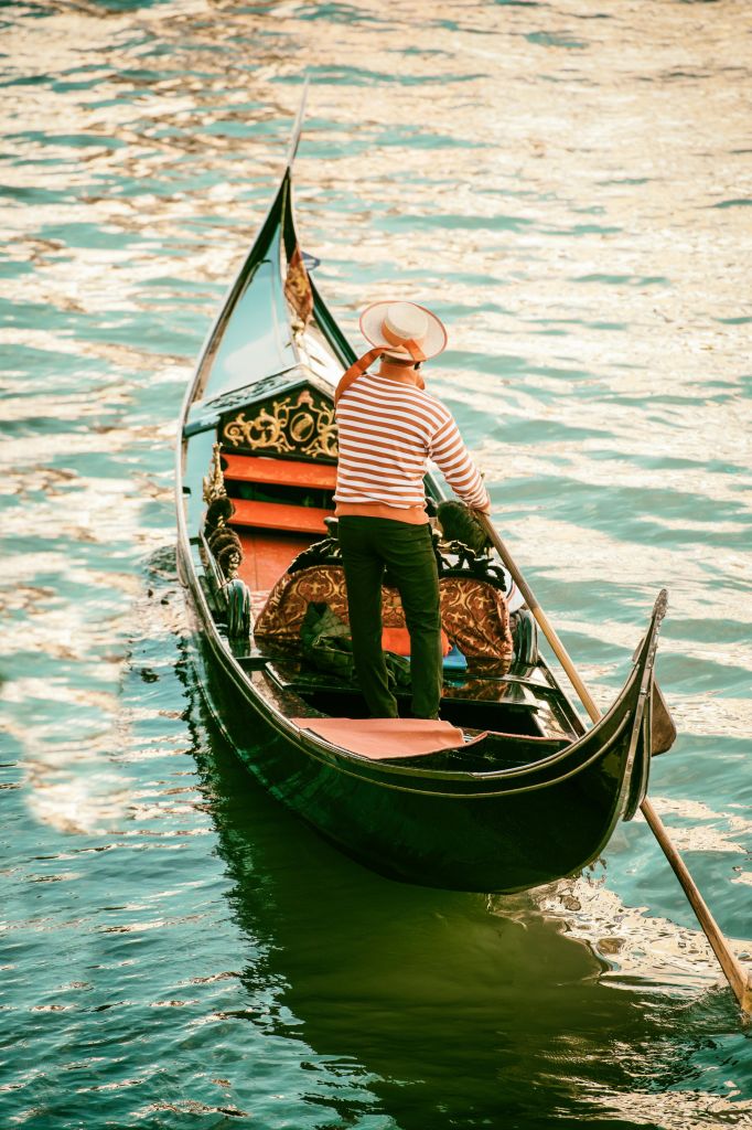 줄무늬 셔츠와 모자를 쓴 곤돌라 선수가 햇빛이 내리쬐는 잔잔한 바다에서 곤돌라를 조종합니다.