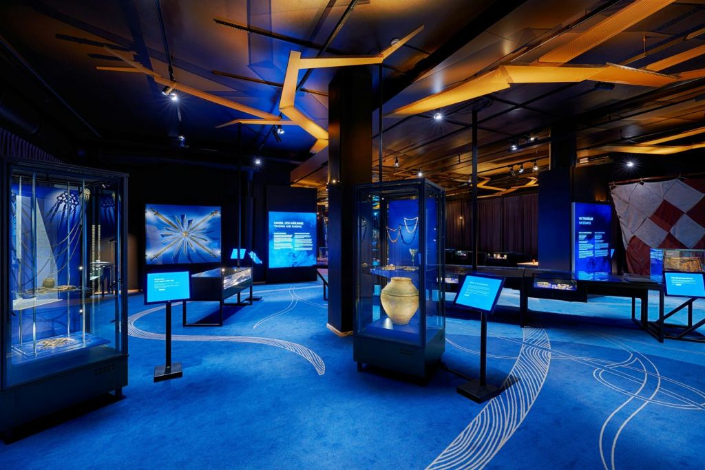 파란색 테마의 조명, 유물이 담긴 진열장, 스탠드의 안내 스크린이 있는 박물관 전시물입니다.