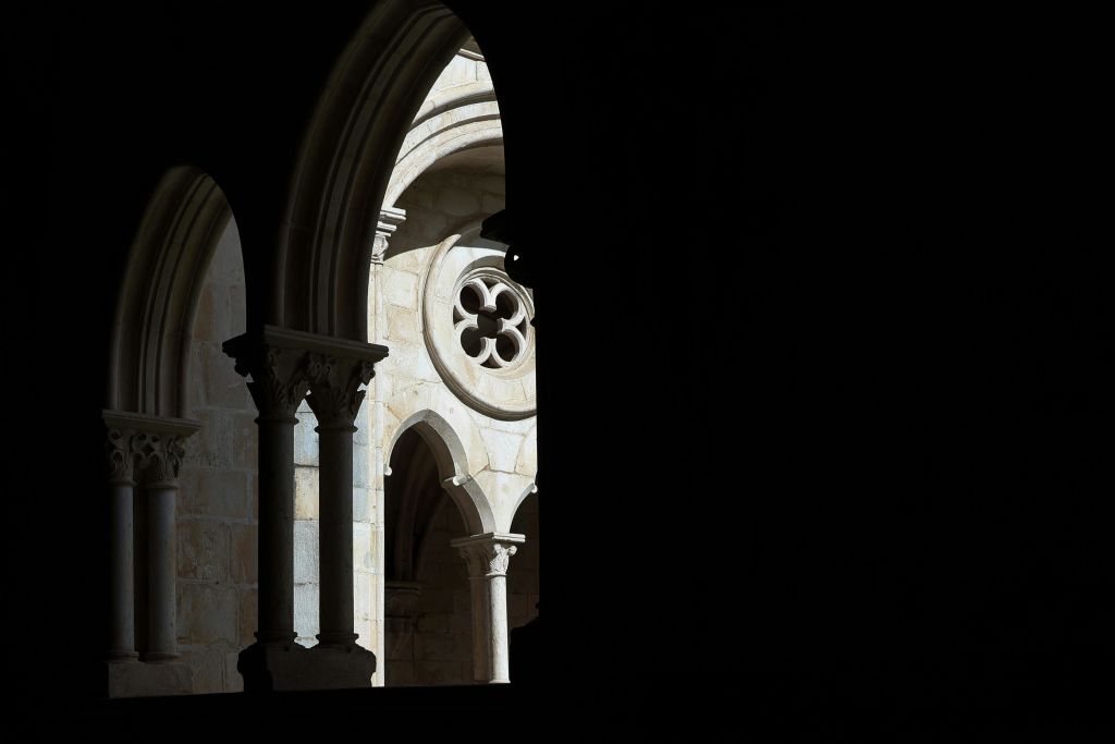 장식용 원형 창문과 기둥이 있는 돌로 된 아치형 통로의 모습으로, 부분적으로 조명이 켜져 있으며 전경에 어두운 그림자가 드리워져 있습니다.