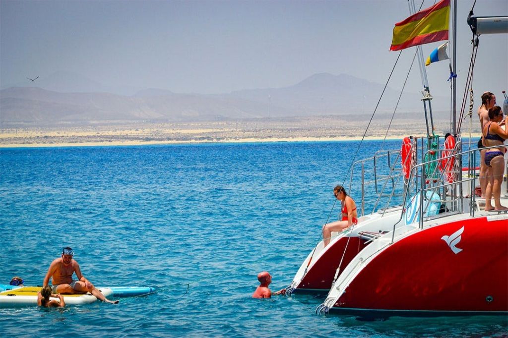산과 맑고 푸른 바다를 배경으로 붉은 선체와 깃발이 달린 보트 근처에서 바다를 즐기는 사람들.