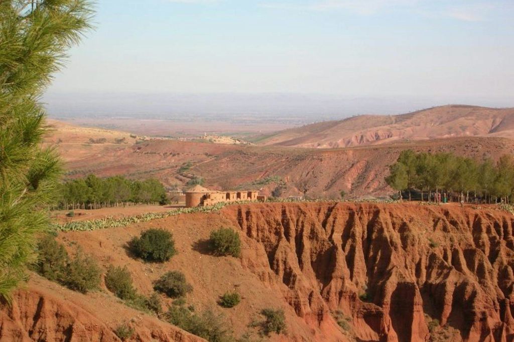 붉은 바위 지형, 희박한 초목, 저 멀리 맑은 하늘 아래 건물들이 어우러진 건조한 풍경.